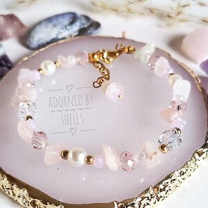 May include: A delicate bracelet featuring pink and white beads, including pearls, crystals, and rose quartz. The bracelet has a gold clasp and is displayed on a pink and gold background. The text "Adorned by Shells" is visible on the background.