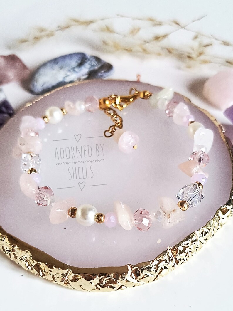 May include: A delicate bracelet featuring pink and white beads, including pearls, crystals, and rose quartz. The bracelet has a gold clasp and is displayed on a pink and gold background. The text "Adorned by Shells" is visible on the background.