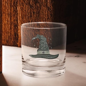 May include: A clear glass tumbler with a green witch's hat design. The hat has a pointed top and a wide brim with a decorative pattern.