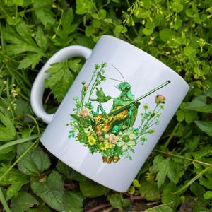 May include: White ceramic mug with a green grasshopper playing a violin surrounded by flowers. The grasshopper is green and has a brown violin. The flowers are white, yellow, and green.