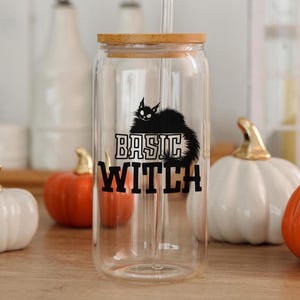 Op de afbeelding: Een heldere glazen beker met een bamboe deksel en rietje. De beker heeft een zwarte kattenafbeelding en de woorden "BASIC WITCH". Decoratieve pompoenen staan op de achtergrond.