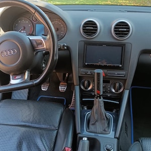Peut inclure: Intérieur de voiture avec tableau de bord et volant. Le volant noir présente des détails argentés et un logo. Le tableau de bord gris comprend des bouches d'aération, un écran et des commandes. Le levier de vitesse est également visible.