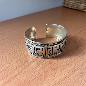 May include: A silver-toned metal cuff bracelet featuring an open design and embossed script characters along the outer band. The bracelet has a polished finish with darkened recessed areas to highlight the intricate lettering and beaded border details.