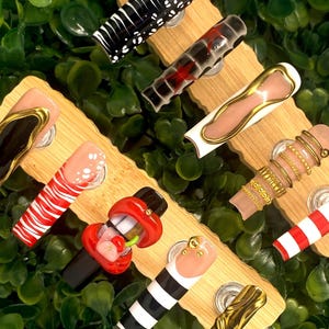 May include: A collection of artificial nails with various designs, including black and white patterns, red and white stripes, and gold accents. Some nails feature intricate details like gold rings and heart shapes. The nails are displayed on wooden stands against a green background.
