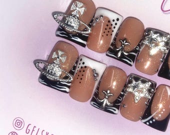 May include: Close-up of a set of false nails with a black and white zebra print design. Each nail is decorated with a silver charm, including a small cross, a star, and a circle with a cross in the center. The nails are on a pink surface with the text "@GELSXGEMSWWW" in the bottom left corner.