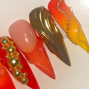 May include: Close-up of five long, pointed artificial nails. The nails feature a variety of designs, including solid red, gold metallic, and gradient orange-yellow. Some nails are adorned with gold embellishments.
