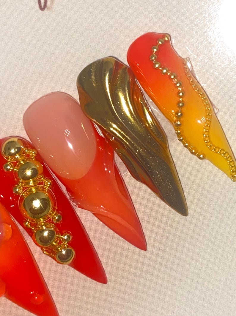May include: Close-up of five long, pointed artificial nails. The nails feature a variety of designs, including solid red, gold metallic, and gradient orange-yellow. Some nails are adorned with gold embellishments.