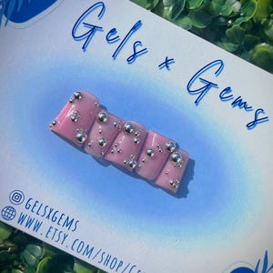 May include: Pink press-on nails with silver 3D embellishments. The nails are displayed on a white card with the text "Gels x Gems" and the social media handle "@gelsxgems".