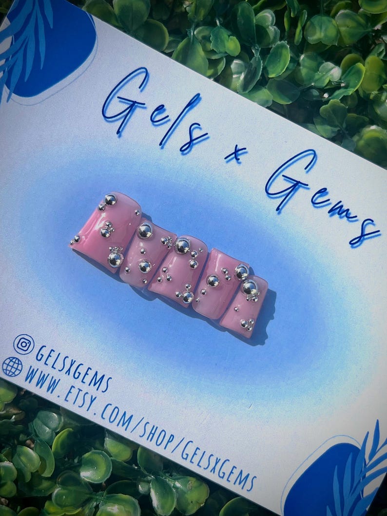May include: Pink press-on nails with silver 3D embellishments. The nails are displayed on a white card with the text "Gels x Gems" and the social media handle "@gelsxgems".