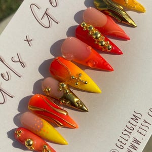 May include: A set of stiletto-shaped false nails in a variety of designs. The nails feature gradients of red, orange, and yellow, with gold accents and embellishments. Some nails have a nude base with gold details.
