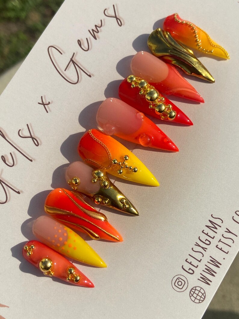 May include: A set of stiletto-shaped false nails in a variety of designs. The nails feature gradients of red, orange, and yellow, with gold accents and embellishments. Some nails have a nude base with gold details.