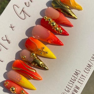 May include: A set of stiletto-shaped artificial nails in various designs. The nails feature a gradient of colors, including yellow, orange, and red, with gold embellishments and a glossy finish. Some nails have a nude base.