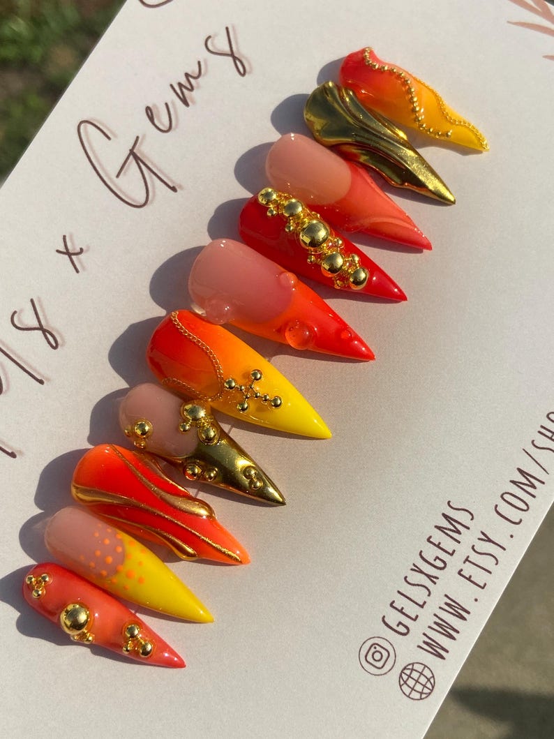 May include: A set of stiletto-shaped artificial nails in various designs. The nails feature a gradient of colors, including yellow, orange, and red, with gold embellishments and a glossy finish. Some nails have a nude base.