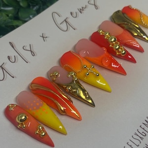 May include: A set of stiletto-shaped press-on nails with a gradient design in shades of red, orange, and yellow. Some nails feature gold embellishments, including studs and cross designs. The text "Gels x Gems" is handwritten on the background.