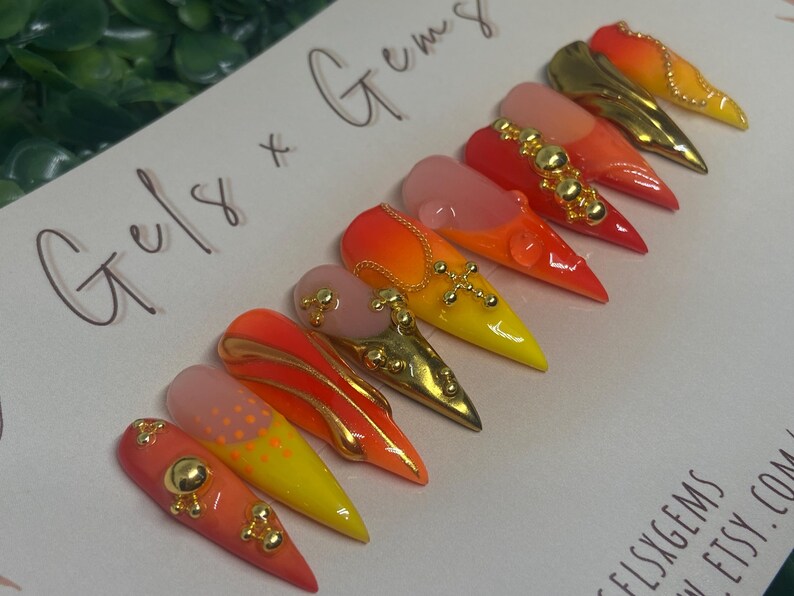 May include: A set of stiletto-shaped press-on nails with a gradient design in shades of red, orange, and yellow. Some nails feature gold embellishments, including studs and cross designs. The text "Gels x Gems" is handwritten on the background.