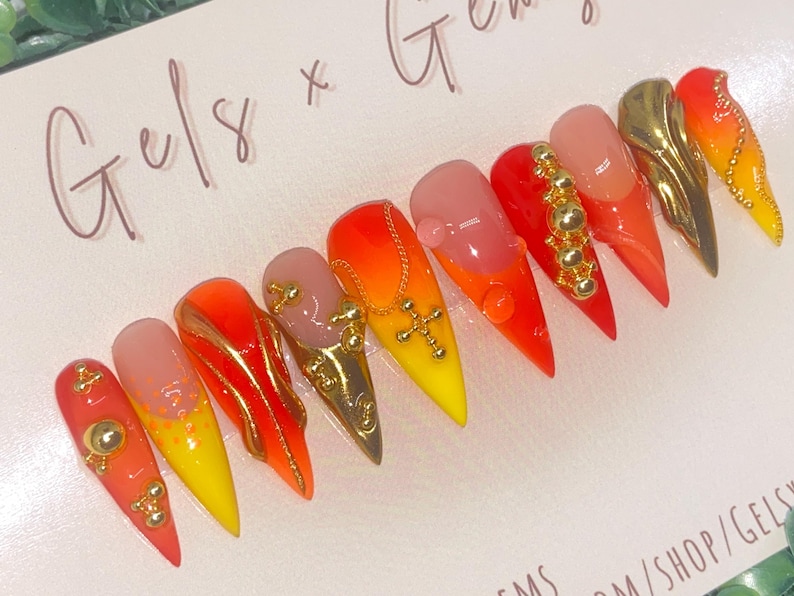 May include: A set of ten stiletto-shaped false nails. Each nail features a gradient design in shades of red, orange, and yellow. Some nails are adorned with gold-colored embellishments, including beads, crosses, and metallic accents. The text "Gels x Gems" is visible.