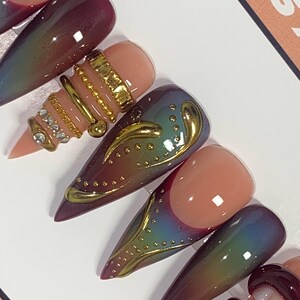 May include: Close-up of several long, pointed artificial nails. The nails feature a variety of designs, including ombre effects, gold embellishments, and intricate patterns. Colors include shades of pink, burgundy, and iridescent hues.