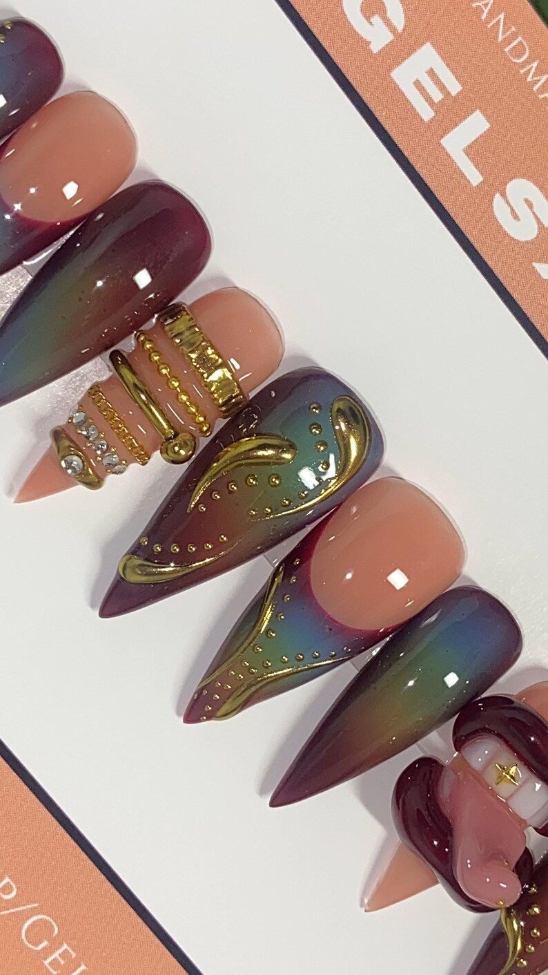 May include: Close-up of several long, pointed artificial nails. The nails feature a variety of designs, including ombre effects, gold embellishments, and intricate patterns. Colors include shades of pink, burgundy, and iridescent hues.