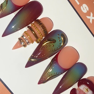 May include: A set of long, pointed false nails with a gradient effect, transitioning from purple to green. Some nails feature gold embellishments and ring designs. The text "GELSX HANDMADE" is visible on a card.