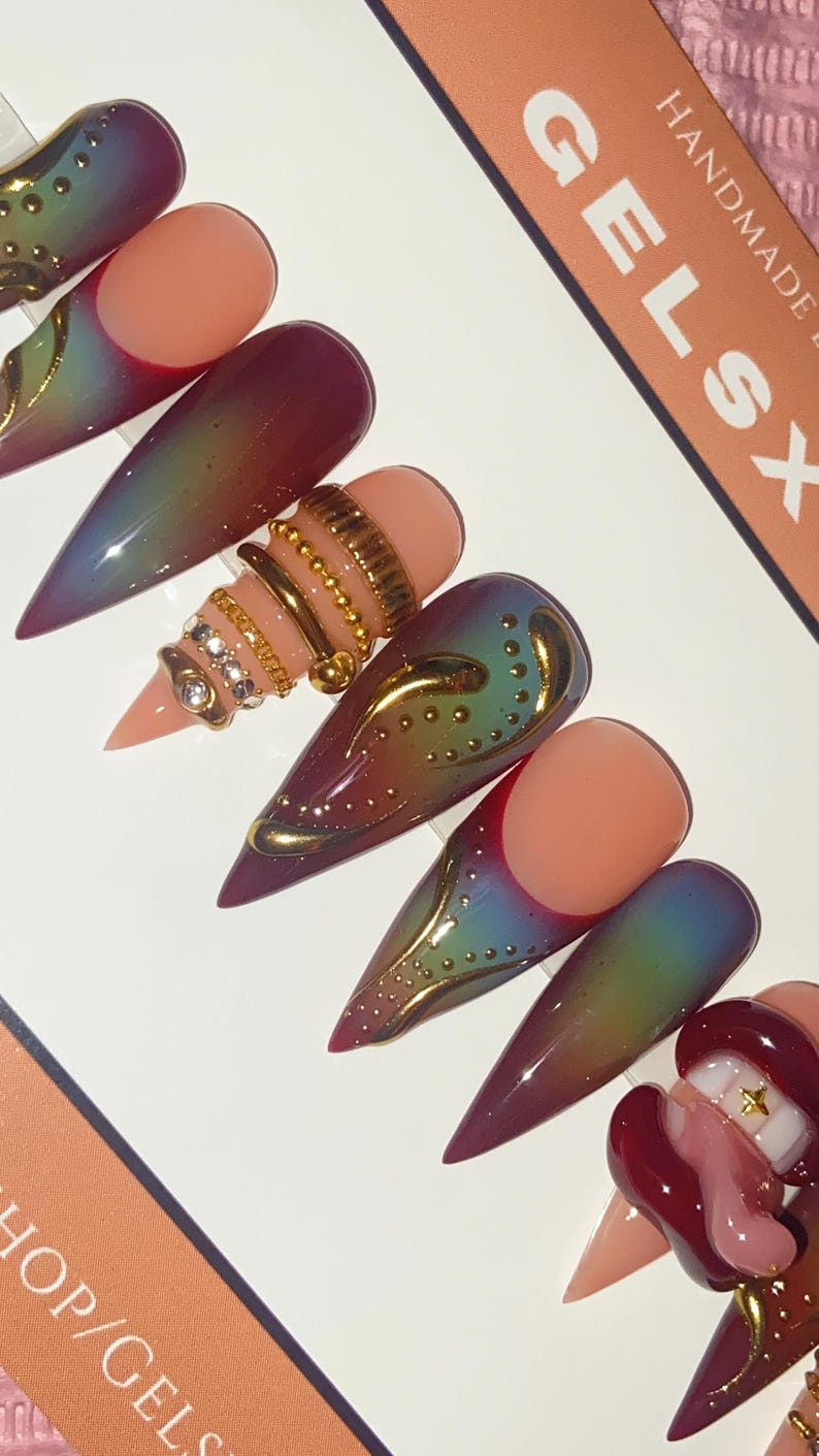 May include: A set of long, pointed false nails with a gradient effect, transitioning from purple to green. Some nails feature gold embellishments and ring designs. The text "GELSX HANDMADE" is visible on a card.