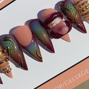 May include: A set of long, pointed press-on nails with various designs. Some nails feature a gradient of colors, gold accents, and a lip design with a gold tooth. The nails are displayed on a white surface.