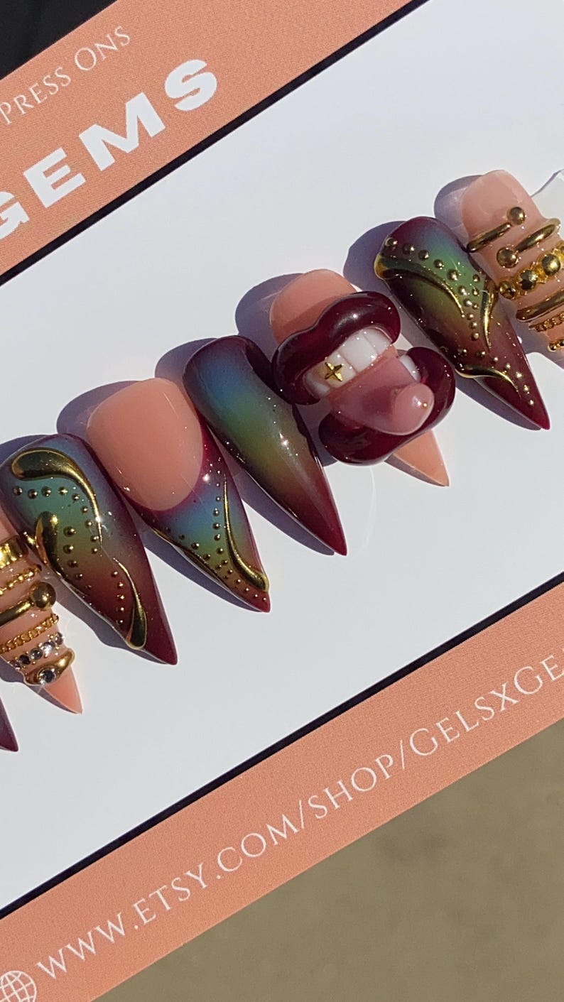 May include: A set of long, pointed press-on nails with various designs. Some nails feature a gradient of colors, gold accents, and a lip design with a gold tooth. The nails are displayed on a white surface.