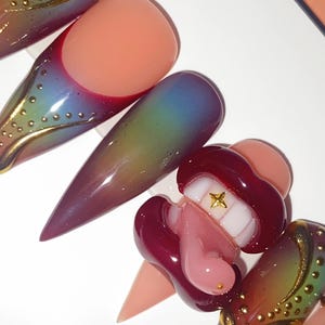 May include: A set of long, pointed artificial nails in various designs. Some nails feature a gradient of colors, while others have gold embellishments and a lip-shaped design with a gold star. The nails are in shades of pink, red, and green.