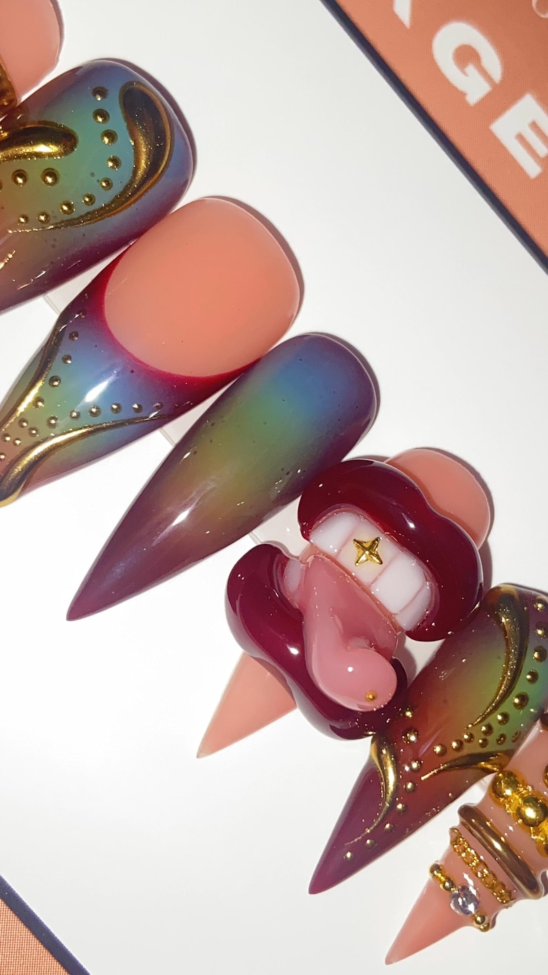 May include: A set of long, pointed artificial nails in various designs. Some nails feature a gradient of colors, while others have gold embellishments and a lip-shaped design with a gold star. The nails are in shades of pink, red, and green.