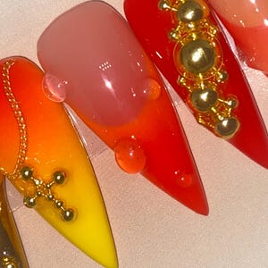 May include: Close-up of several long, pointed acrylic nails. The nails feature a gradient of colors, including yellow, orange, and red. Some nails are adorned with gold-colored charms, including a cross and decorative beads. Clear droplets are also present.