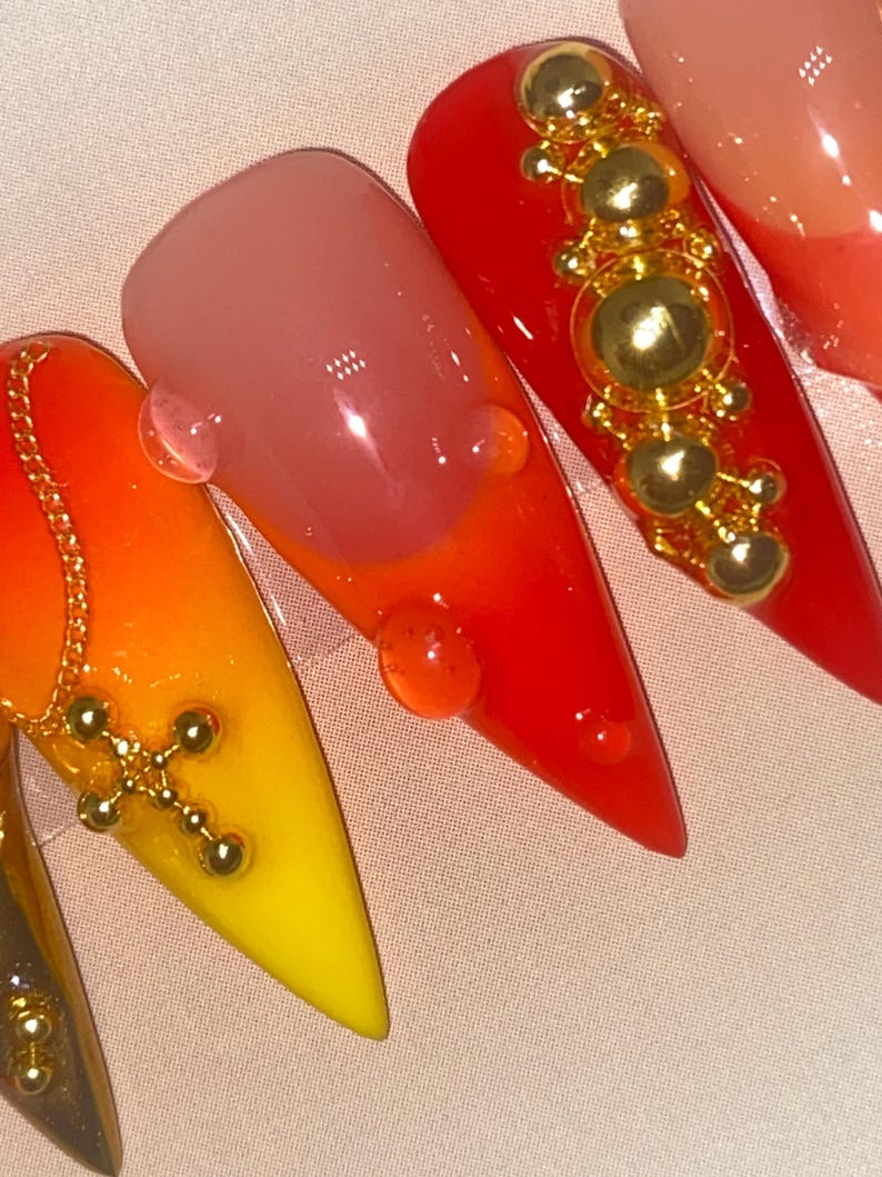 May include: Close-up of several long, pointed acrylic nails. The nails feature a gradient of colors, including yellow, orange, and red. Some nails are adorned with gold-colored charms, including a cross and decorative beads. Clear droplets are also present.