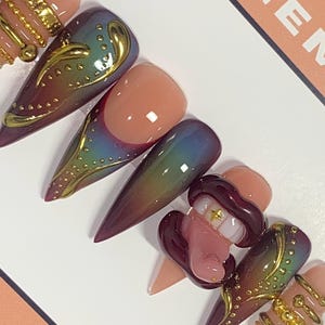 May include: A set of almond-shaped press-on nails with a variety of designs. Some nails feature a gradient of colors, while others have gold embellishments and a 3D lip design with a gold tooth. The nails are arranged on a white surface.