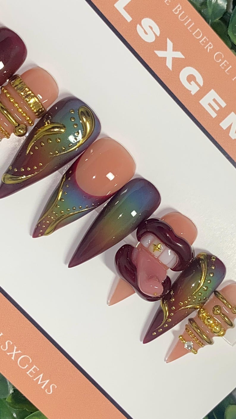 May include: A set of almond-shaped press-on nails with a variety of designs. Some nails feature a gradient of colors, while others have gold embellishments and a 3D lip design with a gold tooth. The nails are arranged on a white surface.