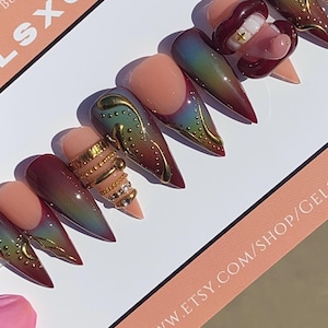 May include: A set of long, pointed press-on nails with a variety of designs. Some nails feature a nude base with gold embellishments, while others have a gradient of colors. One nail has a mouth design with teeth. The text "LSXGEMS" is visible.