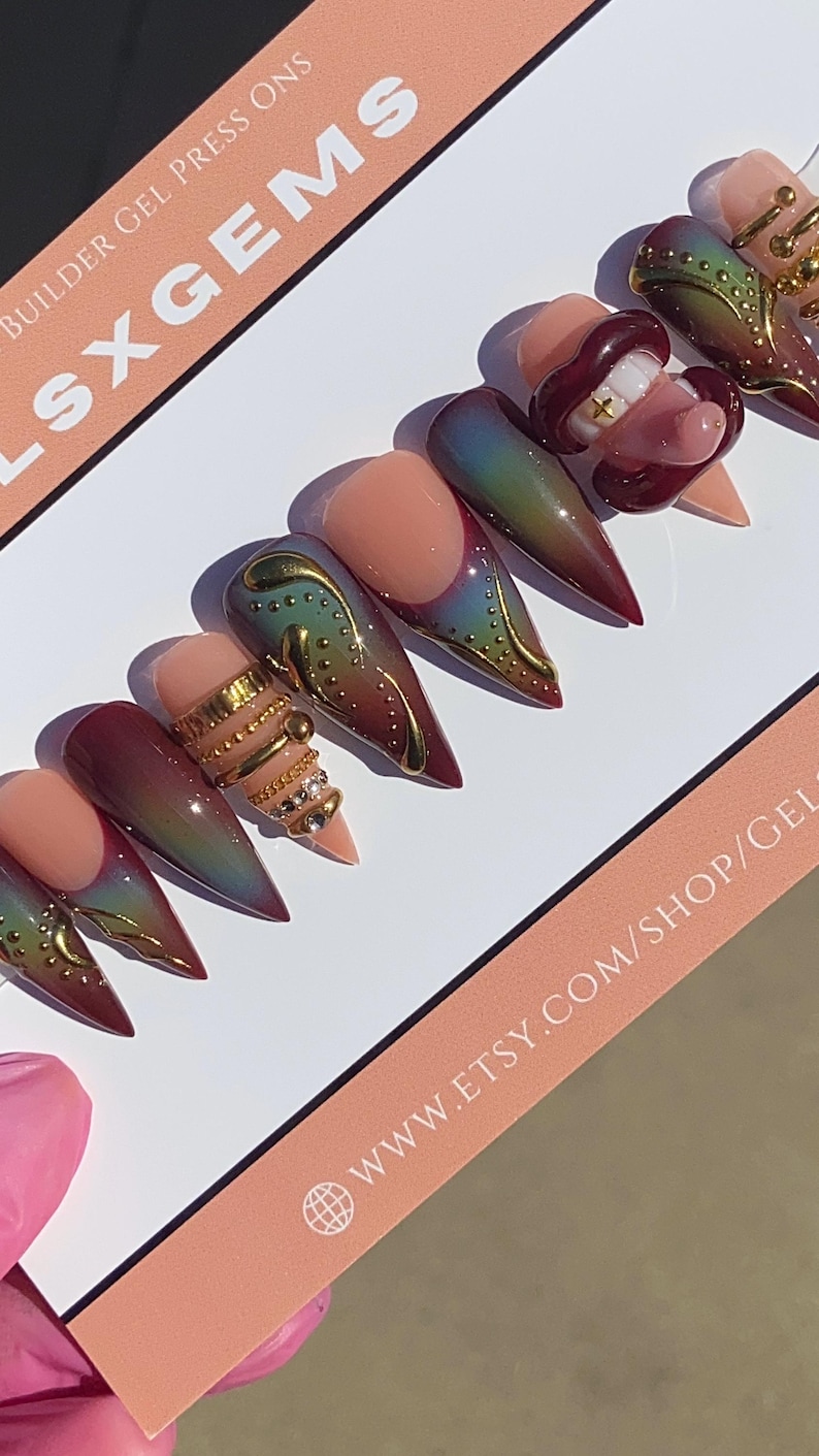 May include: A set of long, pointed press-on nails with a variety of designs. Some nails feature a nude base with gold embellishments, while others have a gradient of colors. One nail has a mouth design with teeth. The text "LSXGEMS" is visible.