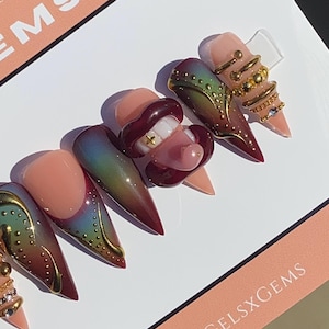 May include: A set of long, almond-shaped artificial nails. The nails feature a gradient design with colors ranging from nude to burgundy, accented with gold details and a 3D mouth design with a gold tooth. The nails are displayed on a white surface.