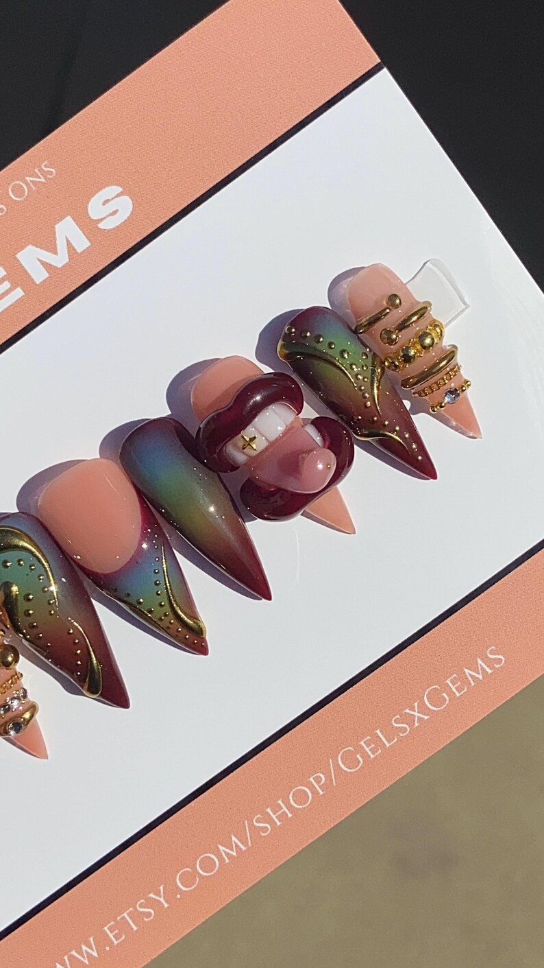 May include: A set of long, almond-shaped artificial nails. The nails feature a gradient design with colors ranging from nude to burgundy, accented with gold details and a 3D mouth design with a gold tooth. The nails are displayed on a white surface.