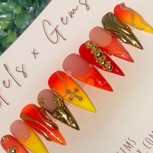 May include: A set of stiletto-shaped false nails with a gradient of colors from pink to yellow, adorned with gold embellishments. Some nails feature gold charms and studs. The text "Gels x Gems" is visible.
