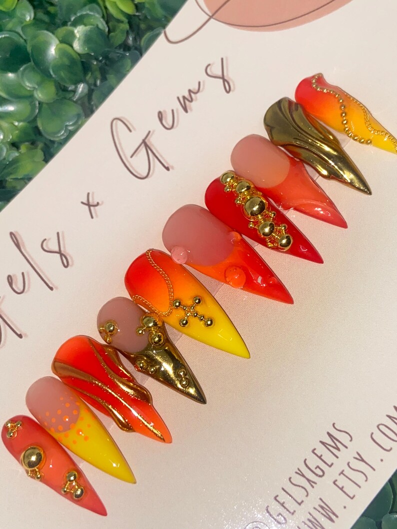 May include: A set of stiletto-shaped false nails with a gradient of colors from pink to yellow, adorned with gold embellishments. Some nails feature gold charms and studs. The text "Gels x Gems" is visible.