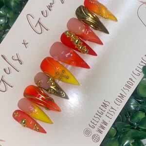 May include: A set of almond-shaped press-on nails with a gradient of colors, including red, orange, and yellow. Some nails feature gold embellishments and gems. The card reads "Gels x Gems" and includes a website and social media handle.