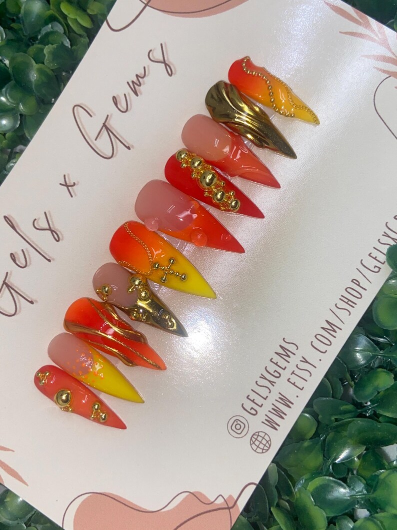 May include: A set of almond-shaped press-on nails with a gradient of colors, including red, orange, and yellow. Some nails feature gold embellishments and gems. The card reads "Gels x Gems" and includes a website and social media handle.