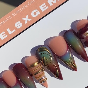 May include: A set of handmade gel press-on nails. The nails feature a gradient design with colors ranging from red to green, accented with gold embellishments and rhinestones. The text "ELSXGEMS" is visible.