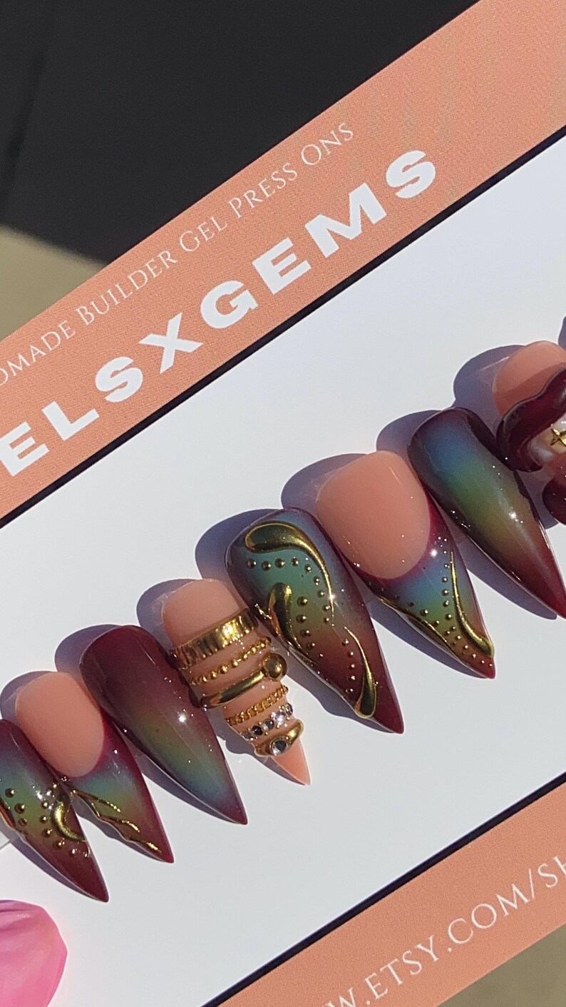 May include: A set of handmade gel press-on nails. The nails feature a gradient design with colors ranging from red to green, accented with gold embellishments and rhinestones. The text "ELSXGEMS" is visible.