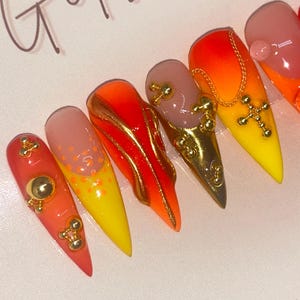 May include: A set of stiletto-shaped artificial nails with a gradient of colors, including red, orange, yellow, and nude. Some nails feature gold embellishments, including charms and a cross pendant. The nails are glossy and appear to be professionally done.