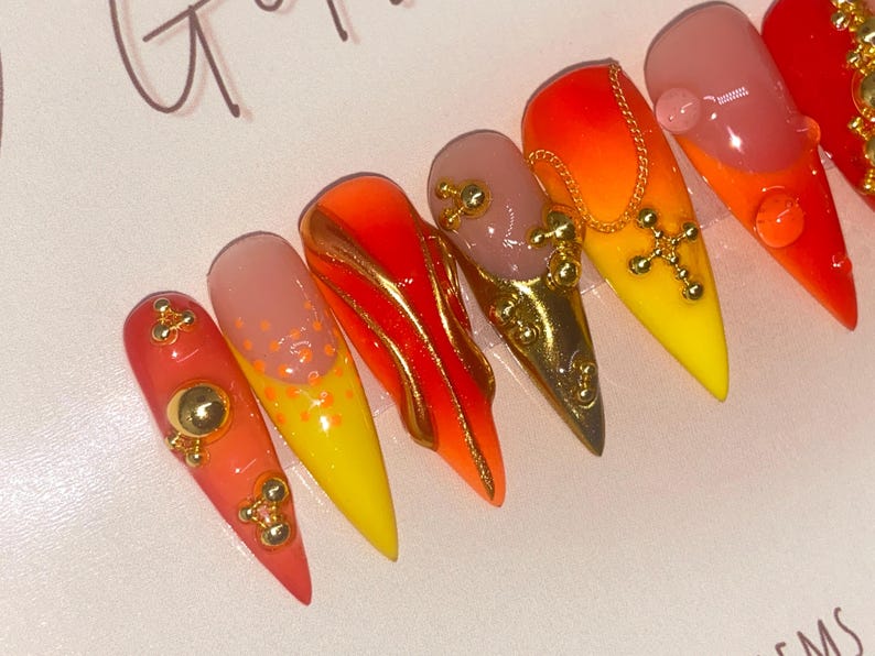 May include: A set of stiletto-shaped artificial nails with a gradient of colors, including red, orange, yellow, and nude. Some nails feature gold embellishments, including charms and a cross pendant. The nails are glossy and appear to be professionally done.