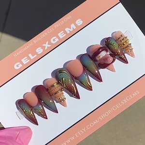 May include: A set of handmade builder gel press-on nails in various designs. The nails feature a gradient of colors, gold accents, and intricate embellishments. The card reads "GELSXGEMS" and "WWW.ETSY.COM/SHOP/GELSXGEMS".
