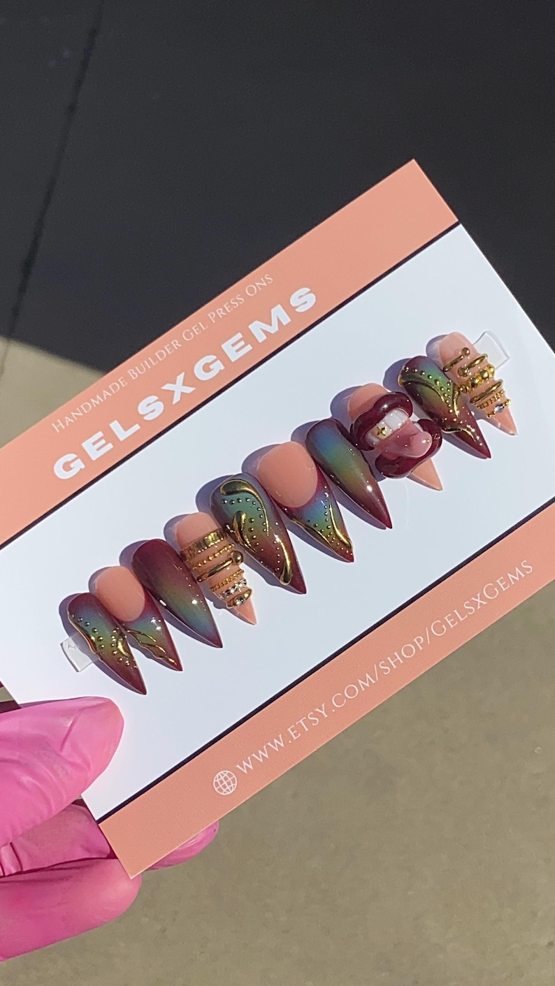 May include: A set of handmade builder gel press-on nails in various designs. The nails feature a gradient of colors, gold accents, and intricate embellishments. The card reads "GELSXGEMS" and "WWW.ETSY.COM/SHOP/GELSXGEMS".