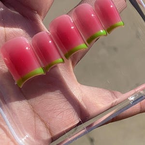 May include: A set of five artificial nails with a watermelon design. The nails are square-shaped with a gradient pink and red color, and a green tip. The nails are held in a clear plastic case.
