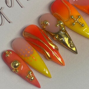 May include: A set of long, almond-shaped artificial nails. The nails feature a gradient of colors, including orange, yellow, and pink. Some nails are adorned with gold embellishments, such as crosses and small spheres. The overall design is vibrant and eye-catching.
