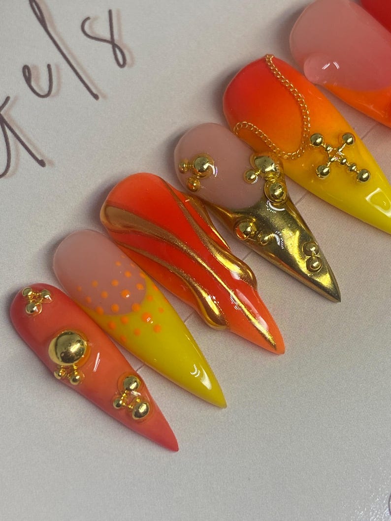 May include: A set of long, almond-shaped artificial nails. The nails feature a gradient of colors, including orange, yellow, and pink. Some nails are adorned with gold embellishments, such as crosses and small spheres. The overall design is vibrant and eye-catching.