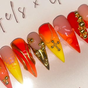 May include: A set of long, pointed artificial nails with a gradient of colors, including yellow, orange, and red. Some nails feature gold embellishments, including beads and a cross pendant. The nails are arranged in a row.